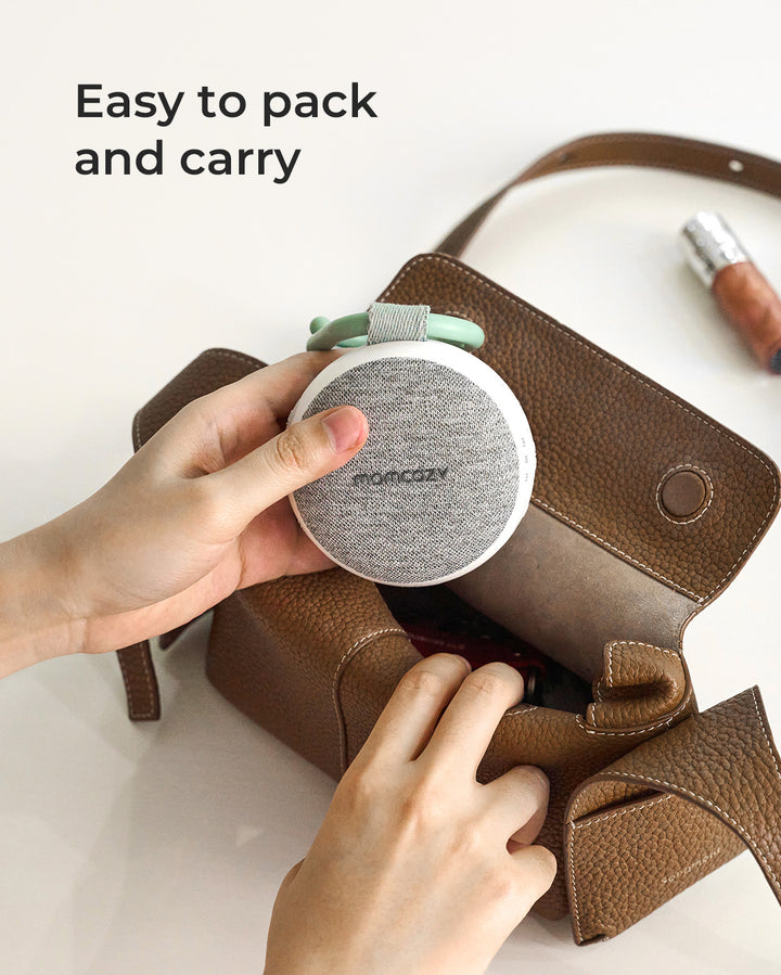 Momcozy portable sound machine, easy to carry in a bag. White speaker, gray fabric, green handle, promoting baby sleep and relaxation.
