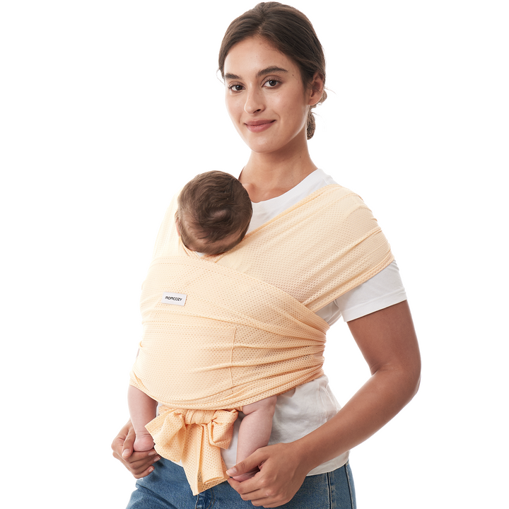 Momcozy baby wrap carrier in peach color, hands-free design for comfortable babywearing. 'Momcozy' logo visible.