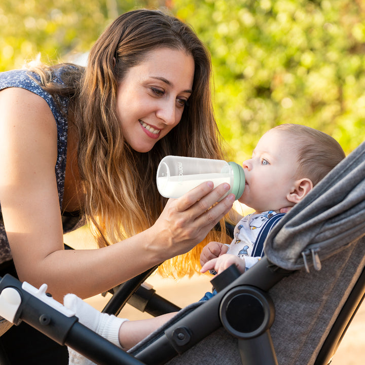 Mother feeding baby from portable bottle outdoors, showcasing Momcozy products.