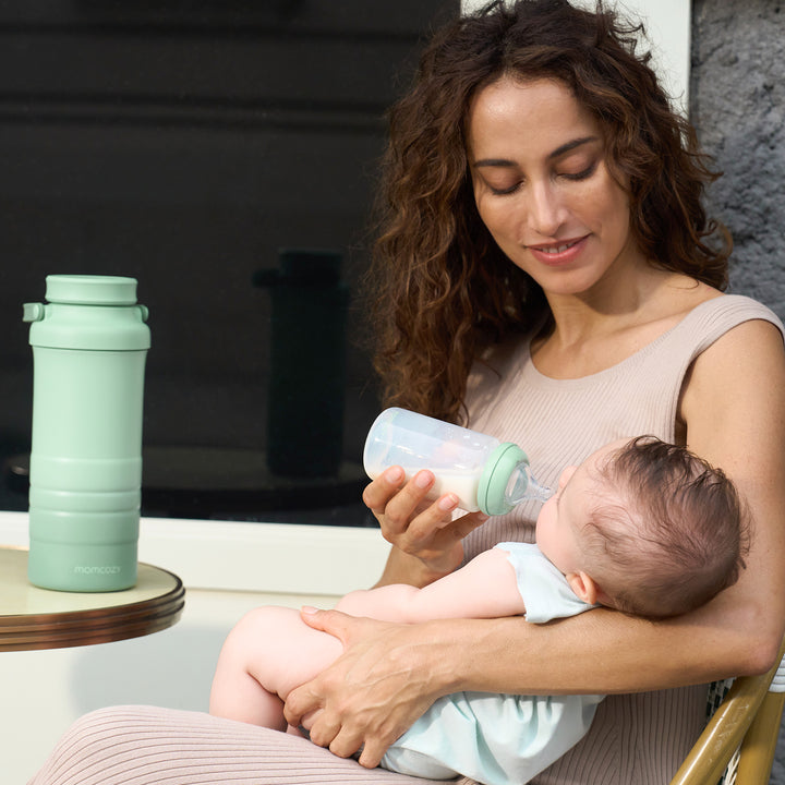 Mom feeding baby from bottle next to green Momcozy breast milk cooler outdoors.