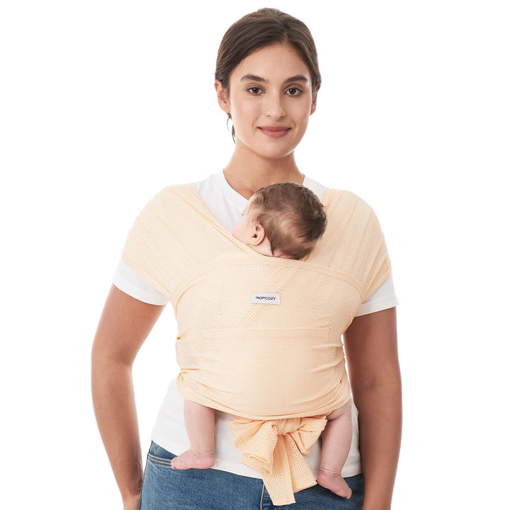 Momcozy baby wrap carrier in peach color, hands-free design for comfortable babywearing. The logo 'Momcozy' is visible.