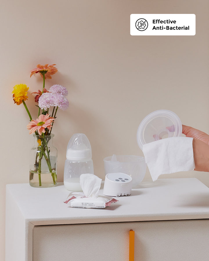 Hand holding a Momcozy wipe above breast pump parts and a bottle, surrounded by flowers.